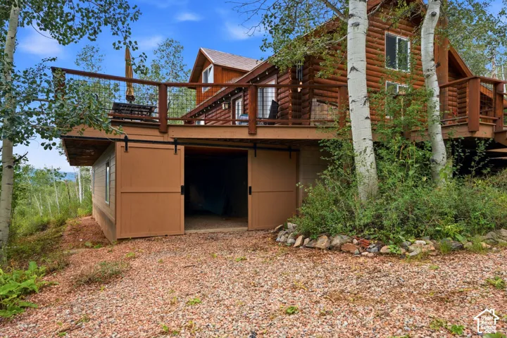 Log cabin exterior with an elevated wood deck featuring a railing, ground-level storage with barn-style sliding doors, and surrounding mature trees