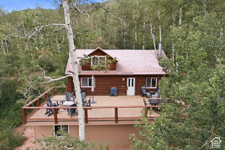 Log-sided exterior with a white entry door, large front deck with dark wood railing and metal infill, dormer window, and a light-colored shingle roof