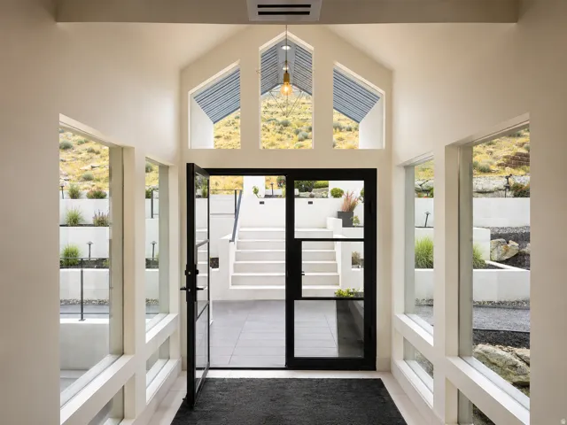 Vaulted entry with transom windows, black-framed glass door, and a multi-tiered outdoor hardscape featuring integrated plantings