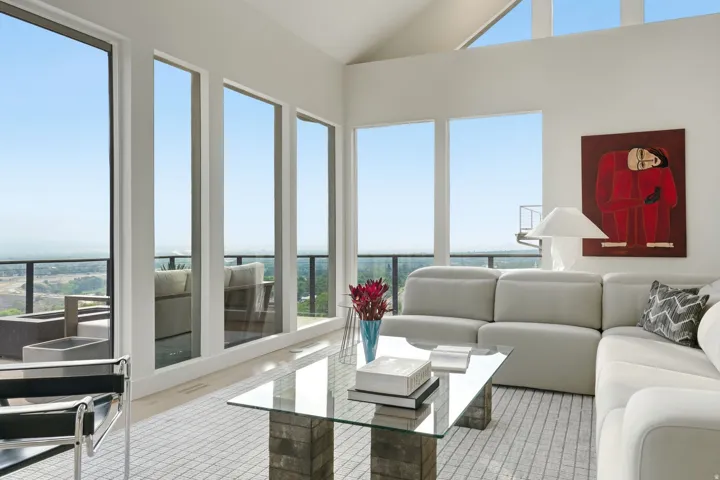 Vaulted ceilings, expansive floor-to-ceiling windows, light wood-finish flooring, and a glass coffee table with stone-finish base