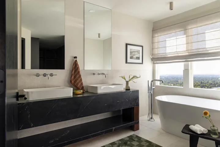 Dual rectangular vessel sinks with wall-mounted faucets, black marble-finish vanity with integrated shelving, freestanding soaking tub with floor-mounted tub filler, and a large window with Roman shades