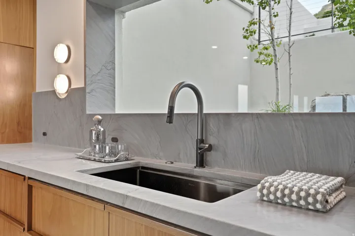 Stone countertop with integrated sink, wood cabinetry, stone backsplash, and wall-mounted lighting