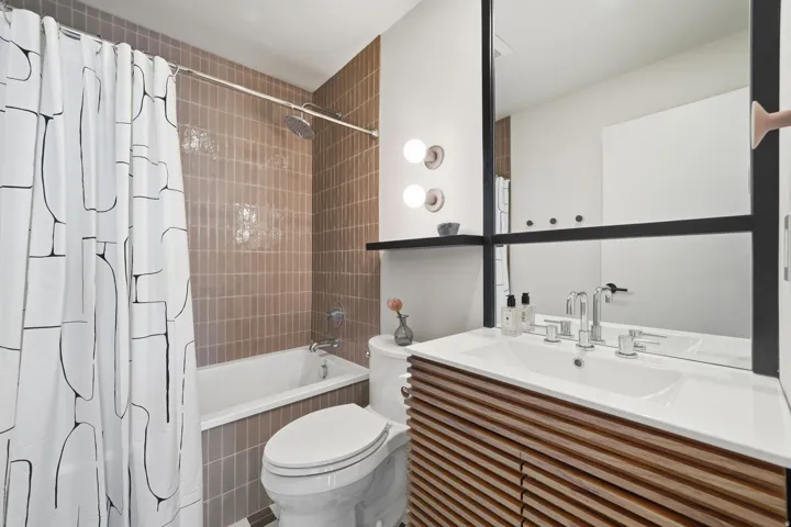 White built-in bathtub with shower, brown vertical tile surround, and a wood-finish vanity with an integrated sink