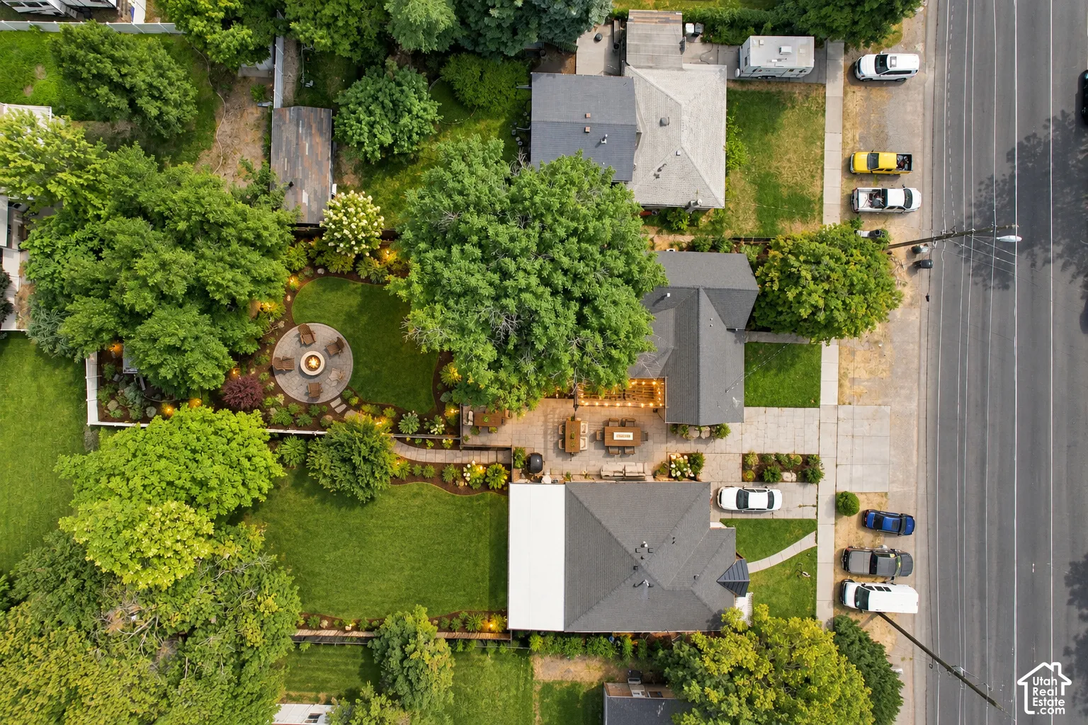 Expansive exterior featuring mature trees, a circular stone patio with a fire pit, a paved patio with string lighting, and manicured green lawns