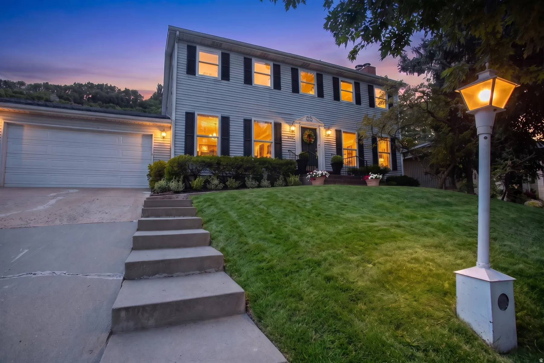Colonial-style residence featuring a two-story facade with blue shutters and illuminated windows, white horizontal siding, and a two-car garage with white paneled doors