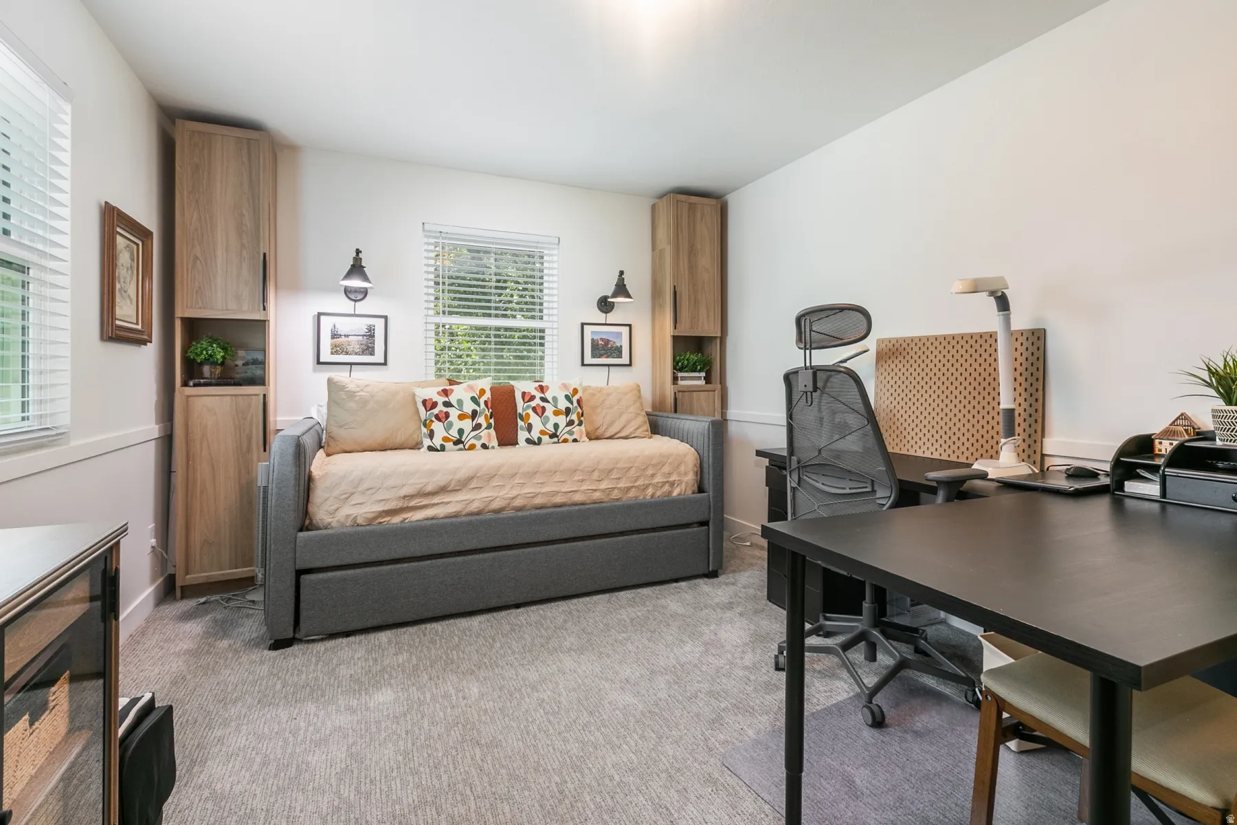 Versatile home office and guest retreat featuring a gray upholstered daybed with storage, black desk workspace with ergonomic seating, light wood-finish built-in cabinetry with shelving, neutral carpet flooring, and abundant natural light from multiple wi