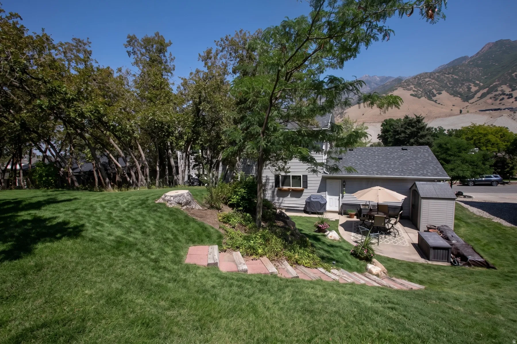 Expansive manicured lawn with mature shade trees and brick hardscaping, backing to hillside terrain