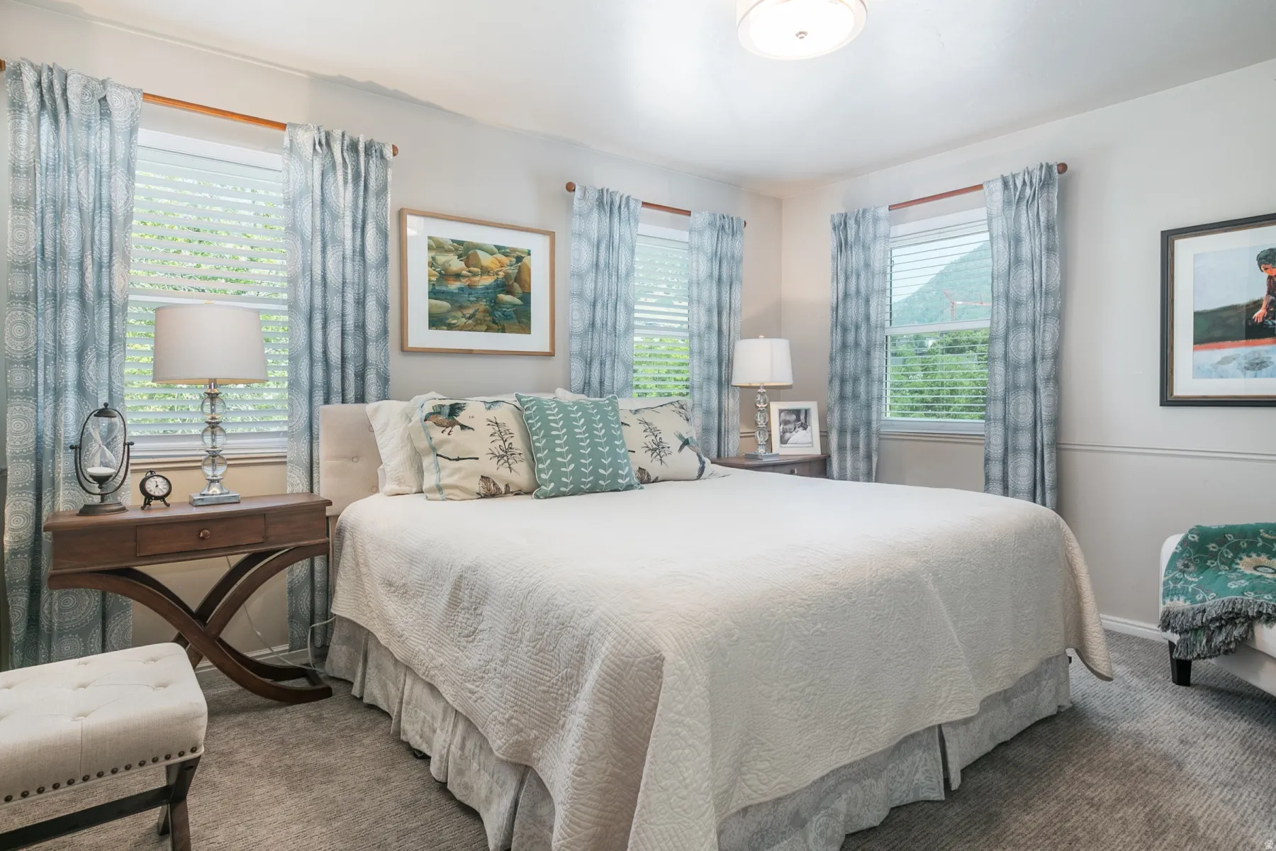 Bright, light-filled bedroom featuring multiple windows with blue patterned curtains, hardwood nightstands with traditional X-frame design, neutral bedding, recessed ceiling lighting, and neutral carpet flooring