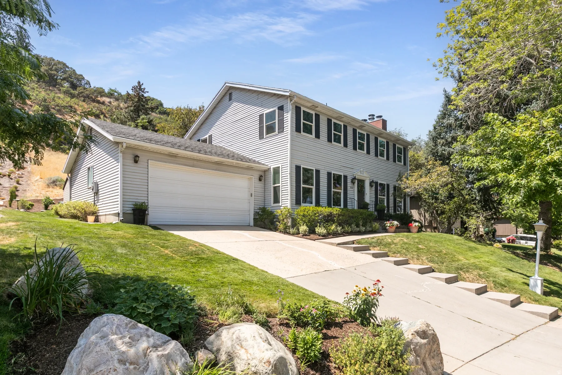 Two-story colonial with white vinyl siding and dark green shutters, featuring a two-car garage, concrete driveway, and manicured front lawn bordered by mature landscaping and natural stone accents