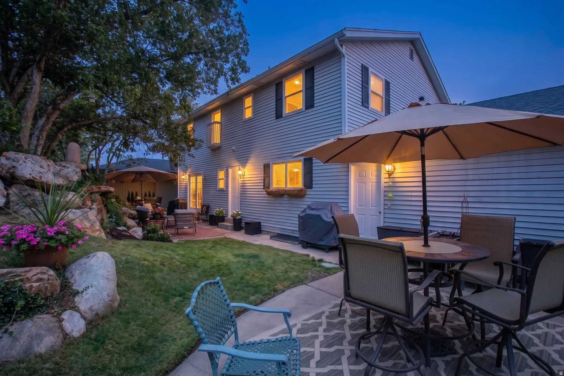 Twilight Entertaining Oasis: This expertly designed outdoor living space features a mature tree canopy, manicured lawn with stone edging, and a covered patio anchored by a large cantilever umbrella and dining table