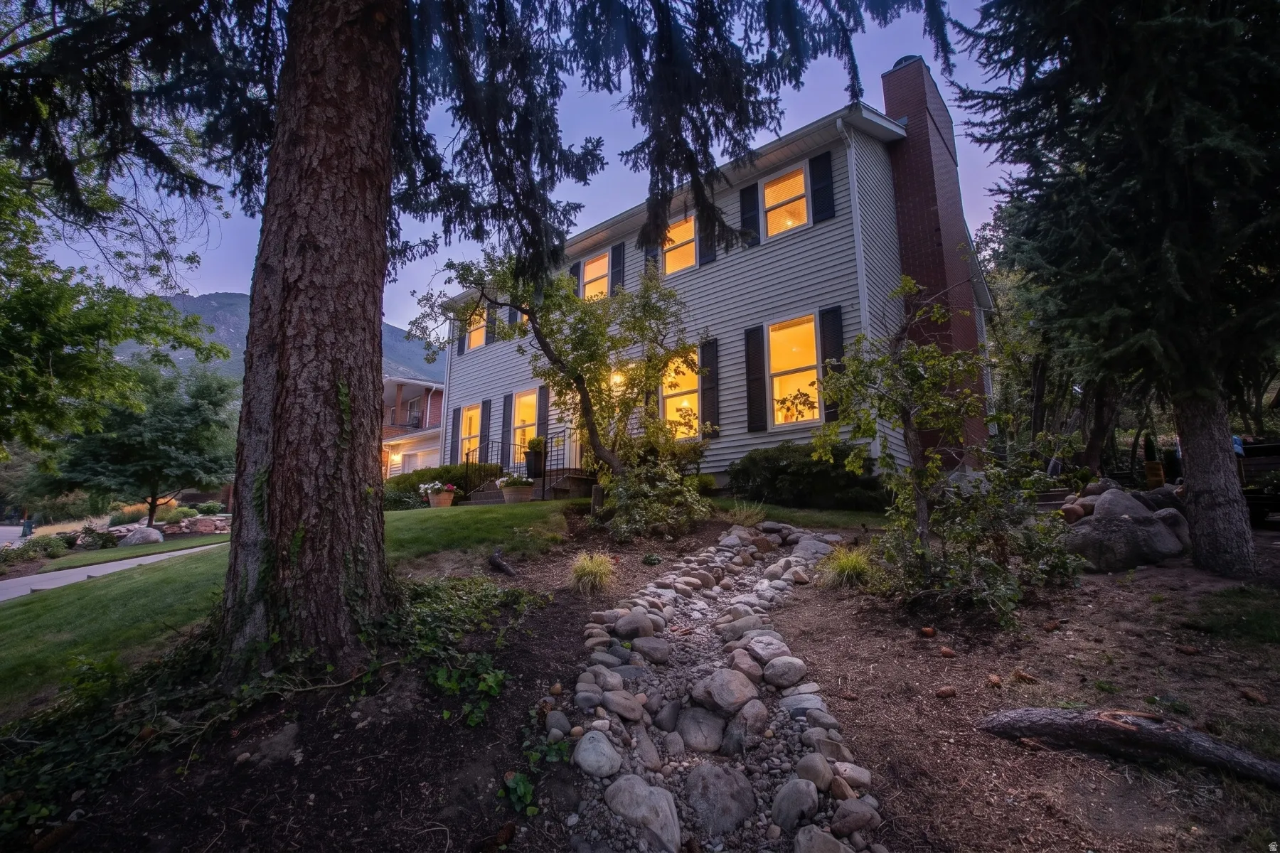 Elegant Colonial residence illuminated by warm interior lighting, nestled among mature evergreen and deciduous trees