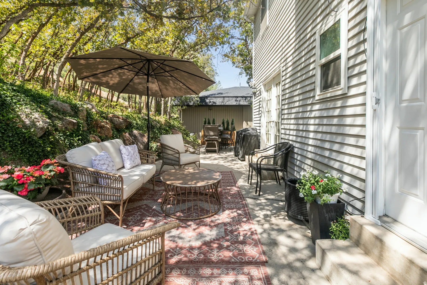 Charming brick patio with mature tree canopy providing dappled shade, woven lounge seating, and decorative planters with flowering shrubs