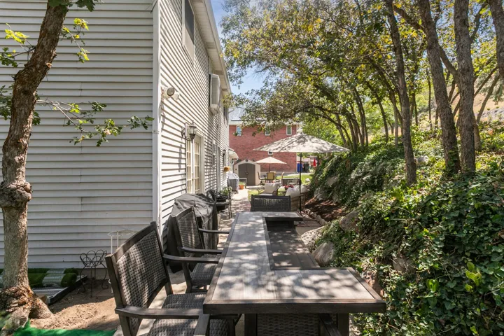 Shaded backyard retreat featuring a composite deck with dining table seating, mature tree canopy providing dappled sunlight, and lush landscaping with hedges and perennial plantings