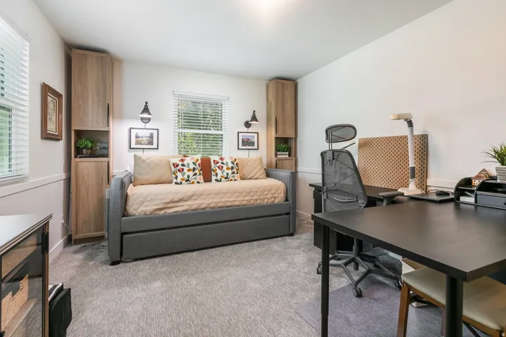 Versatile home office and guest retreat featuring a gray upholstered daybed with storage, black desk workspace with ergonomic seating, light wood-finish built-in cabinetry with shelving, neutral carpet flooring, and abundant natural light from multiple wi