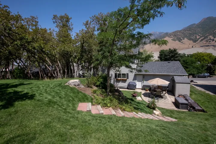 Expansive manicured lawn with mature shade trees and brick hardscaping, backing to hillside terrain