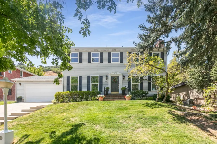 Two-story residence featuring light-colored siding, black window shutters, a two-car garage, and a sloped front lawn