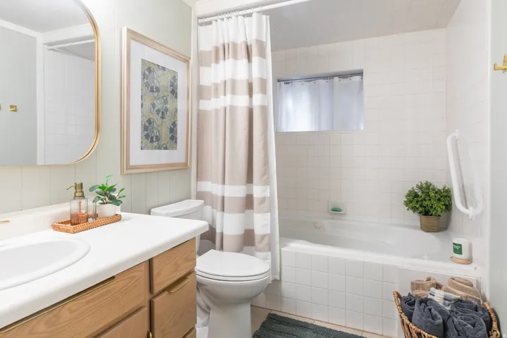 Built-in bathtub with white tile surround, wood-finish vanity with countertop and integrated sink, wall-mounted mirror with gold-tone frame