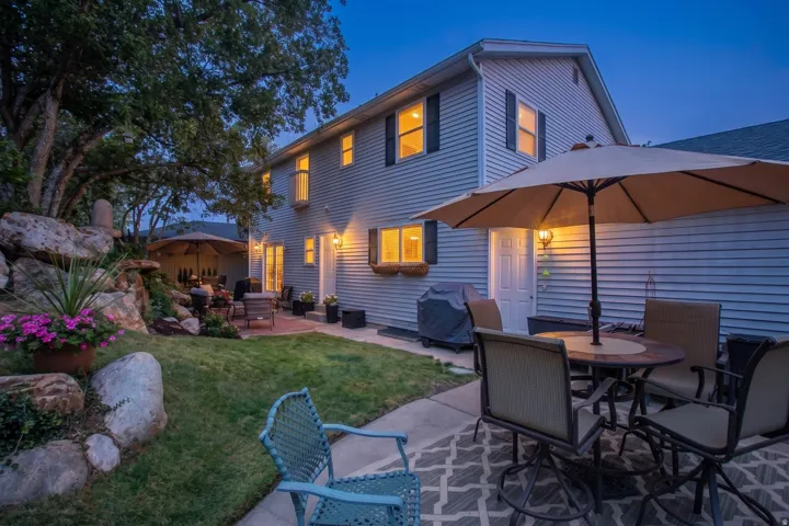 Twilight Entertaining Oasis: This expertly designed outdoor living space features a mature tree canopy, manicured lawn with stone edging, and a covered patio anchored by a large cantilever umbrella and dining table