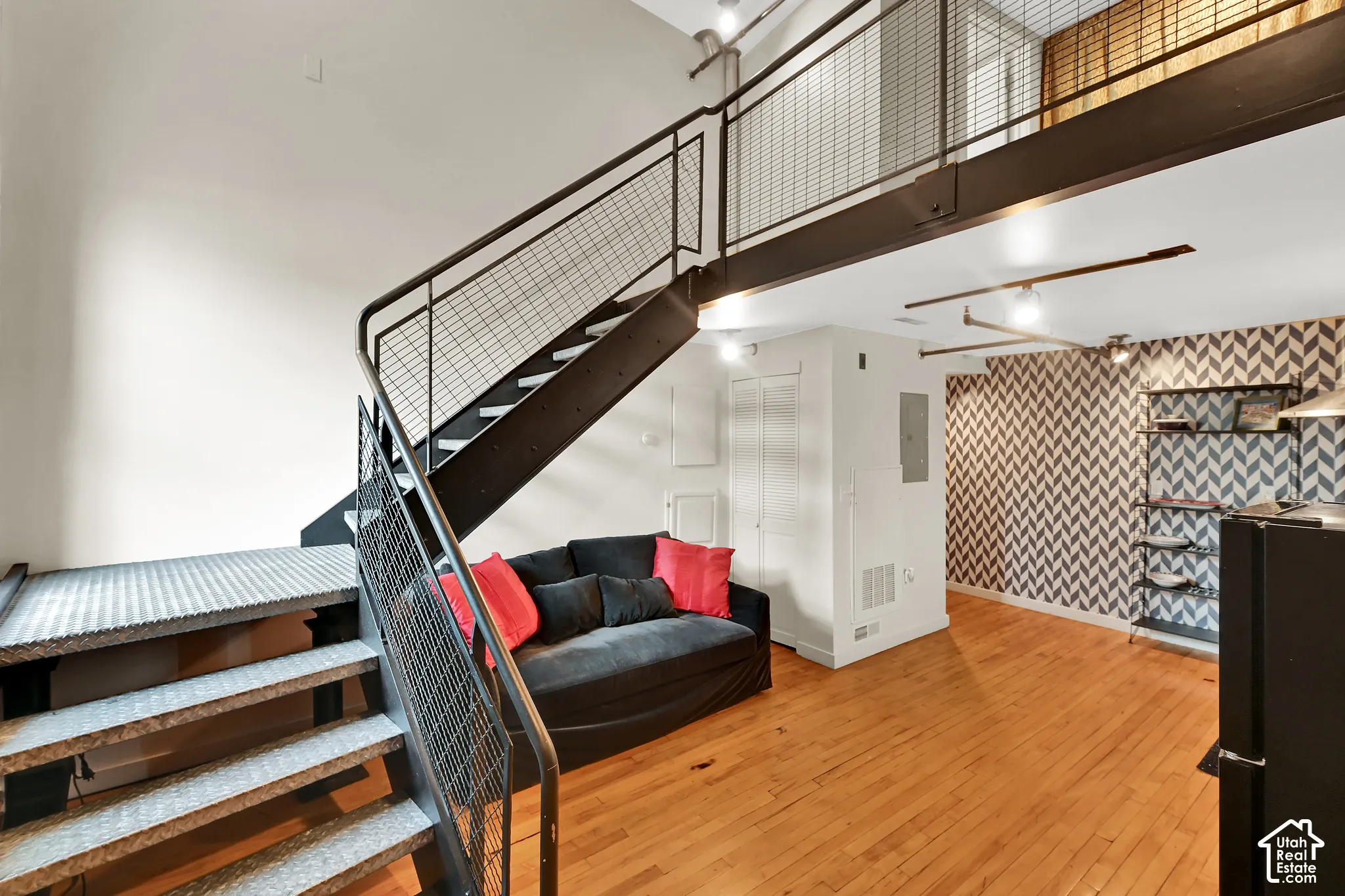 Modern loft-style interior featuring an open floor plan with warm wood-finish flooring, a steel-railed mezzanine staircase with metal mesh panels, and soaring ceilings