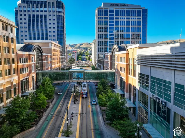 Urban streetscape featuring a tree-lined pedestrian corridor with multiple lanes of paved roadway, modern mixed-use buildings with glass and metal facades, and mountain backdrop visible in the distance under clear skies