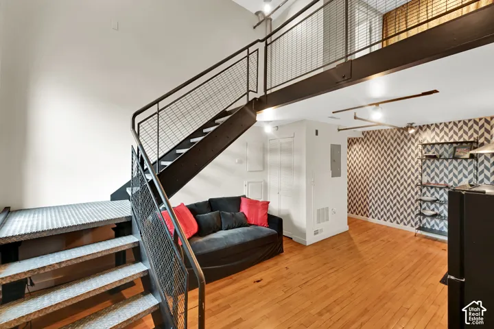 Modern loft-style interior featuring an open floor plan with warm wood-finish flooring, a steel-railed mezzanine staircase with metal mesh panels, and soaring ceilings