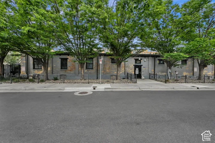 Stone-clad facade with multi-pane windows and black metal fencing, shaded by mature trees lining the streetfront