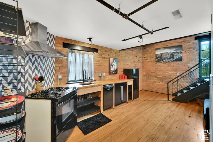 Exposed brick walls, wood-finish flooring, stainless steel range hood, black appliances, and a straight-run staircase with metal railing