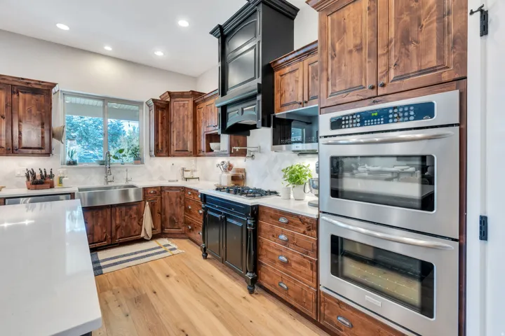 Contemporary kitchen featuring wood cabinetry, stainless steel double ovens, a gas range with a black hood, a stainless steel farmhouse sink.