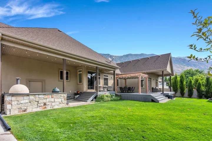 Expansive green lawn, a covered patio with string lighting, a stone-faced outdoor kitchen with a built-in oven, and an additional covered deck with a pergola.