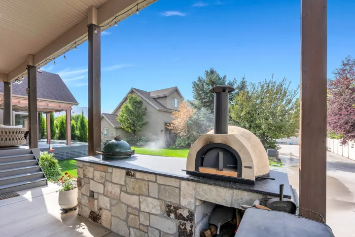 Covered patio featuring substantial stone columns and a wood-finish ceiling, anchoring a custom wood-fired pizza oven with a cream-colored dome and black metal base.