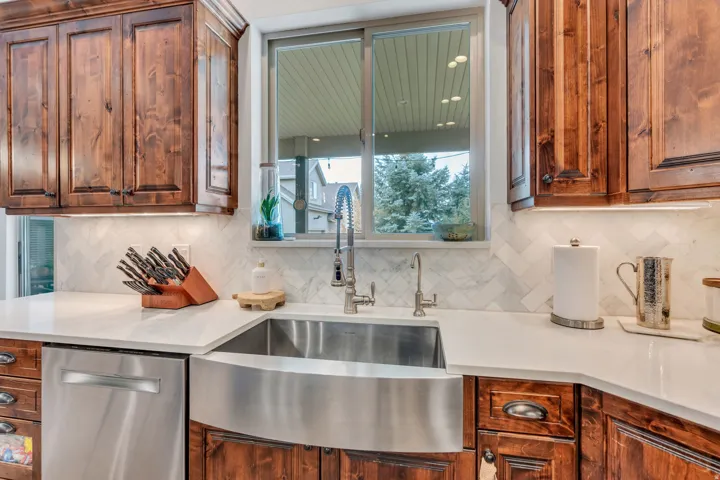Elegant kitchen featuring custom wood cabinetry, a stainless steel farmhouse sink with polished faucet, white custom tile backsplash, and white quartz countertops.