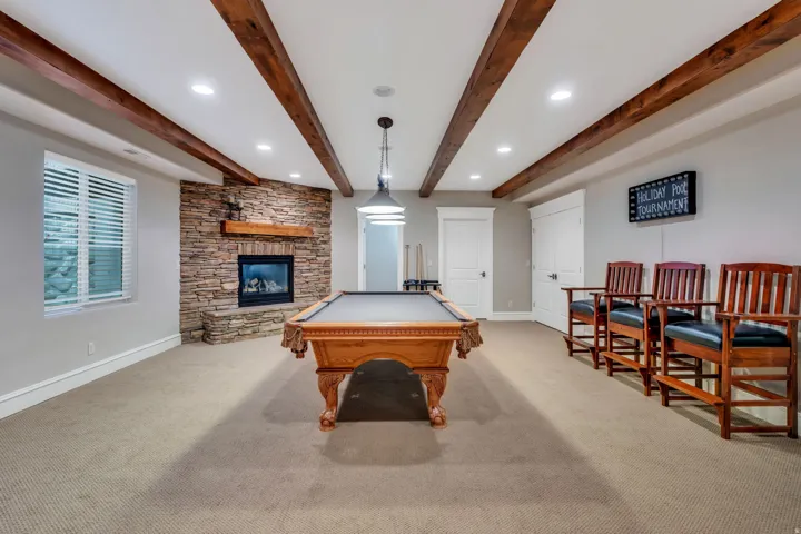Expansive recreation room featuring exposed wood beam ceiling with recessed lighting, brick fireplace with hearth, carpet flooring, and large windows with plantation shutters.