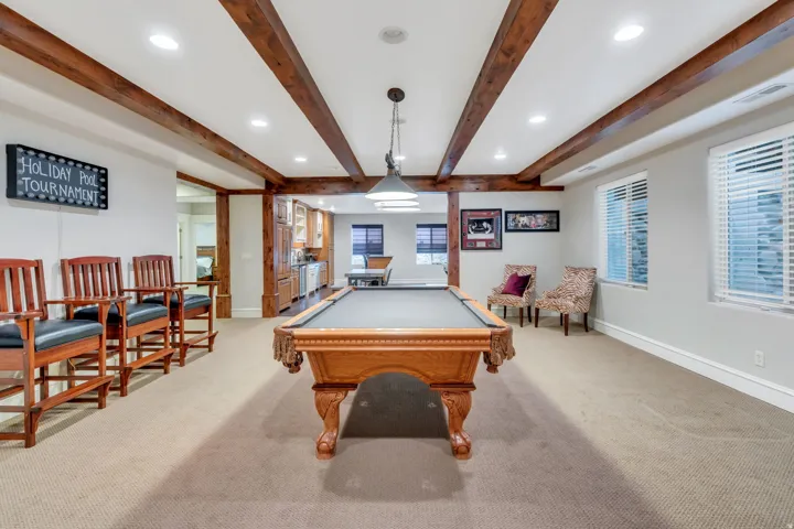Expansive recreation room showcasing exposed wood beam ceiling with recessed lighting, neutral carpet flooring, and abundant natural light from multiple windows.