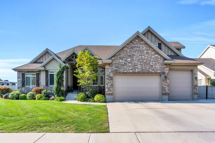 Stunning stone and brick exterior with a three-car garage, manicured landscaping featuring mature shrubs and lush lawn, and elegant architectural detailing including peaked rooflines and decorative stonework accents throughout the façade.