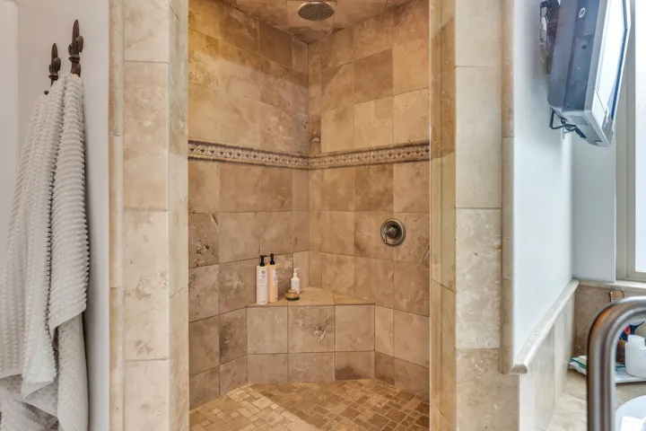 Walk-in shower featuring natural stone-look tile, decorative accent band, integrated corner bench, and mosaic tile flooring.  Three shower heads.