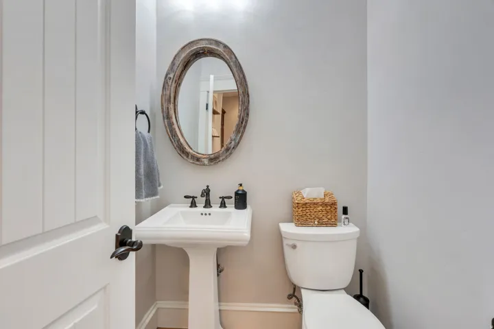 Main floor half guest bath. Pedestal sink with black fixture, toilet, and an oval mirror with a decorative frame.