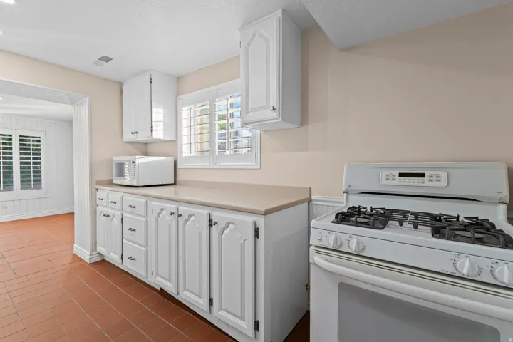 White cabinetry with decorative fronts, light-toned countertops, ceramic tile flooring, gas range, and louvered window treatments