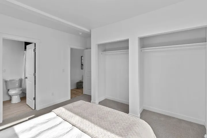 Carpeted room with white walls, two open closet spaces with hanging rods, a visible bathroom with a toilet, and wood-finish flooring in the bathroom