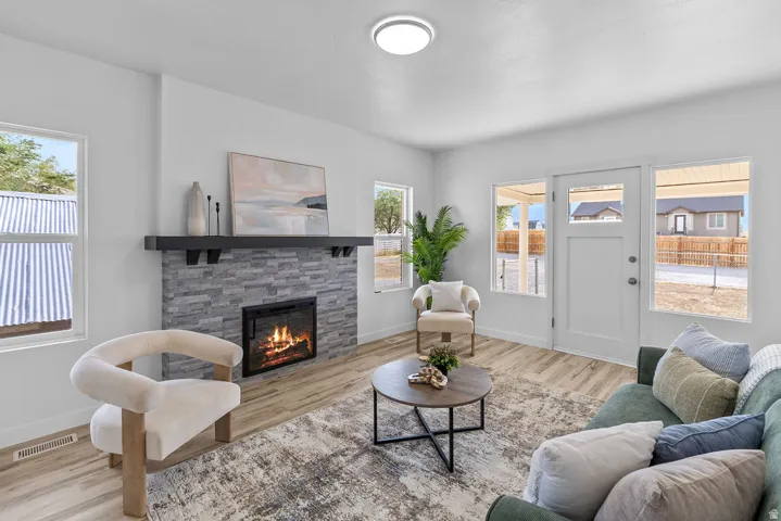 Living space with light wood-finish flooring, a stone-front fireplace with a dark mantel, multiple windows, and a white entry door with glass inserts