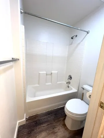 White bathtub with an integrated shower, chrome fixtures, white tile surround, and wood-finish flooring