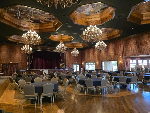Ball room with Hardwood flooring, decorative wall paneling, elevated stage with full-height curtains, multiple ornate chandeliers, and wall sconces