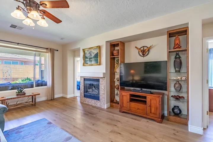 Wood-finish flooring, built-in shelving, fireplace with stone surround, ceiling fan with light fixture, and large window