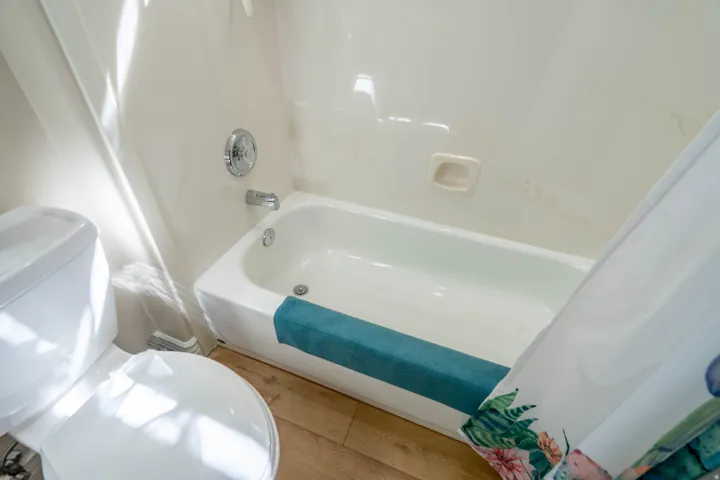 White bathtub with chrome fixtures and light-toned cultured marble wall surround, complemented by wood-finish flooring and a white toilet