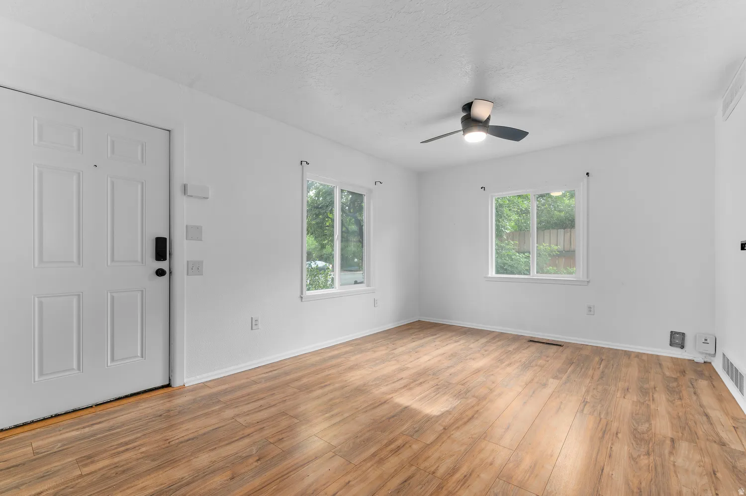 Wood-finish flooring throughout, white paneled entry door, two windows with white trim, and a ceiling fan with integrated lighting