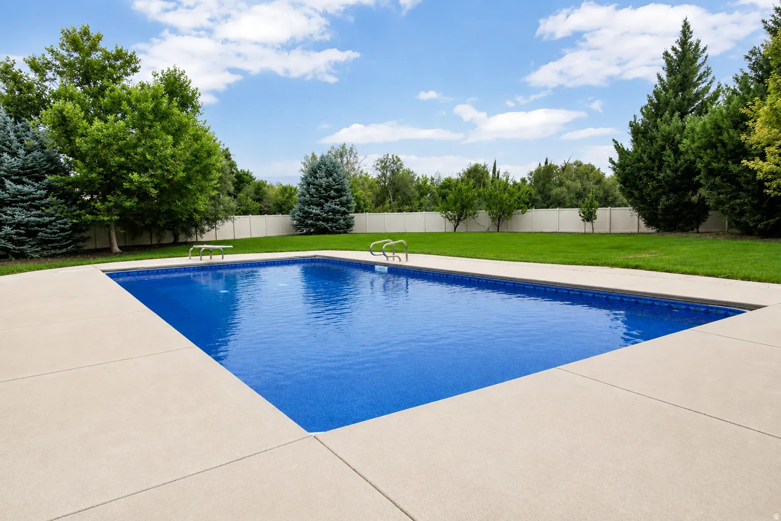 Rectangular inground pool with concrete surround, lush green lawn, mature trees, and privacy fencing