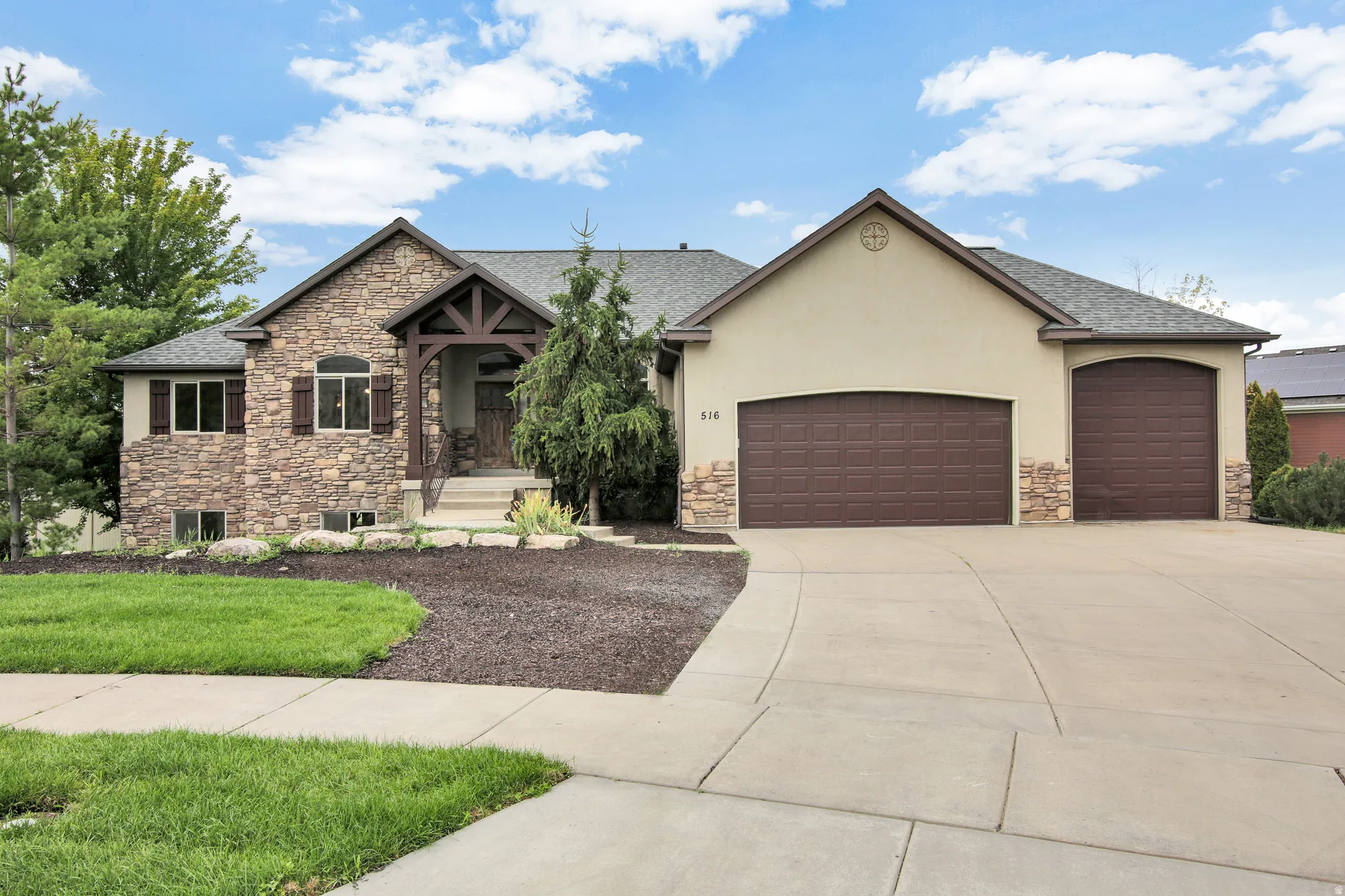 Stone and stucco exterior, covered entry with wood beam accents, multiple-car garage, concrete driveway, and established front landscaping