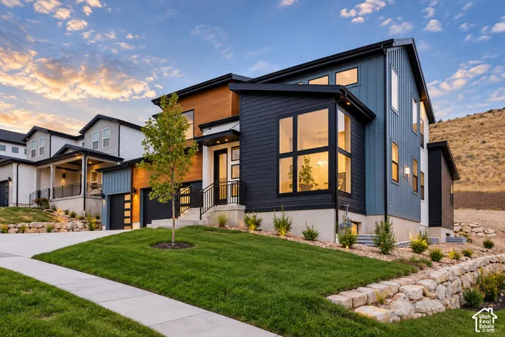 Modern architectural design featuring varied siding materials, expansive windows, a two-car garage, and a landscaped front yard with a stone retaining wall. Image enhanced with AI to show lights on inside.  Back yard is now finished.