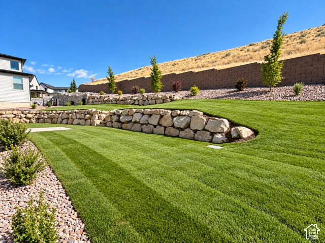 Expansive backyard featuring a manicured lawn, multiple-tiered rock retaining walls, a concrete patio area, and drought-tolerant landscaping