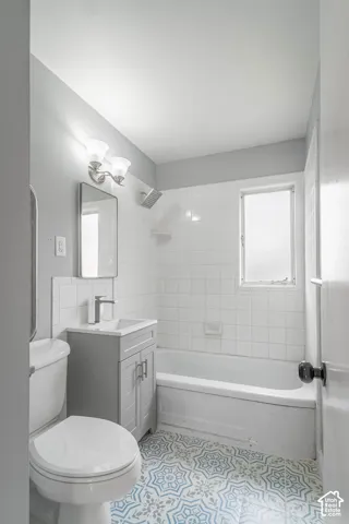 Bathroom featuring patterned tile flooring, a gray vanity cabinet with a rectangular basin, a tiled bathtub surround, and a wall-mounted two-light fixture