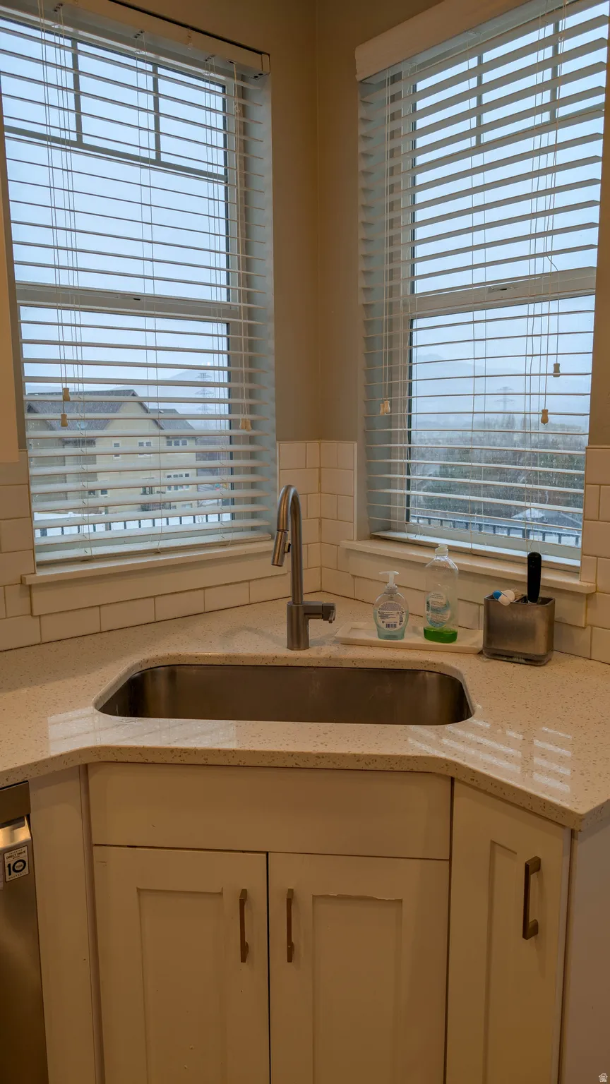 Corner kitchen sink with a stainless steel basin, brushed nickel faucet, light-toned countertop, white subway tile backsplash, and white cabinetry